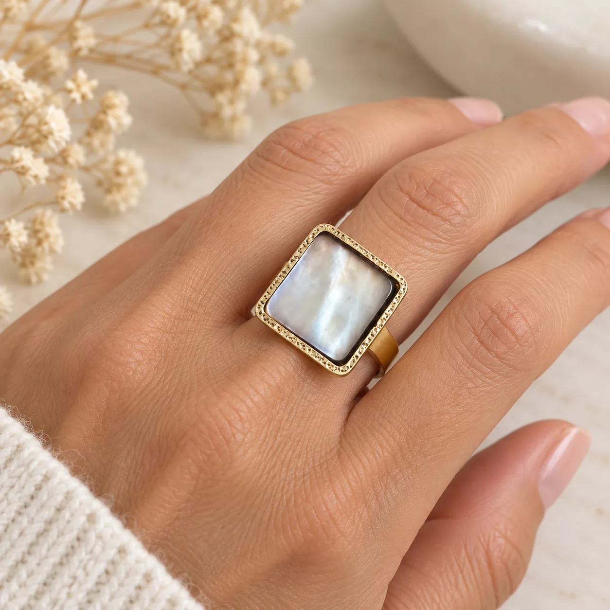 Bague carrée en nacre blanche aux reflets irisés, avec contour doré texturé, portée au doigt